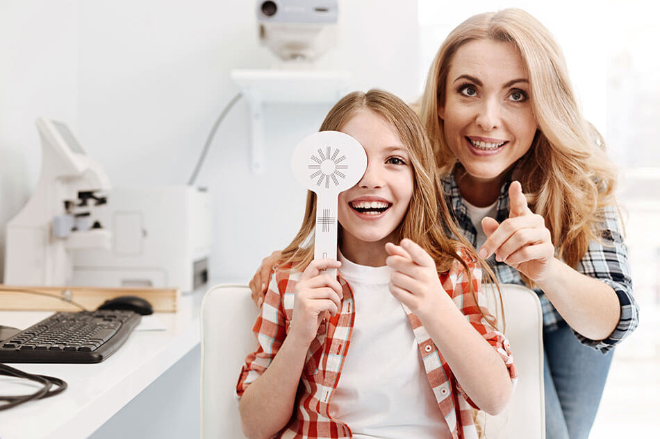 girl smiling with eye paddle along with her mother during eye exam